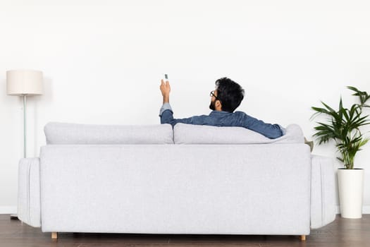 Rear view man sitting on sofa at home, holding remote