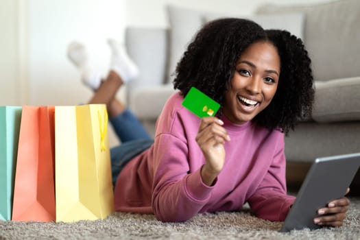 Glad millennial african american curly woman shopaholic lying on floor with bags with purchases recommends credit card