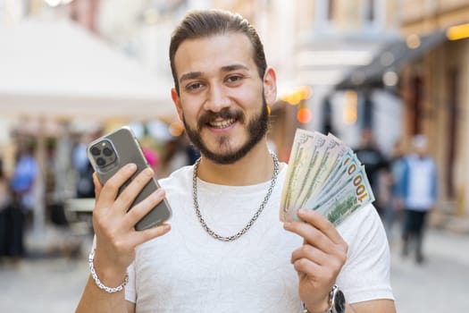Happy young man counting money dollar cash, use smartphone calculator app in urban city street