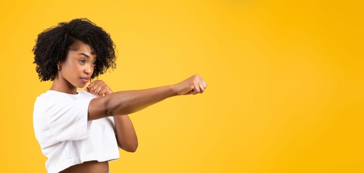 Serious confident millennial african american curly woman in white t-shirt fighting, punching, isolated on yellow studio background, panorama. Self-defense, battle, strength, power, ad and offer