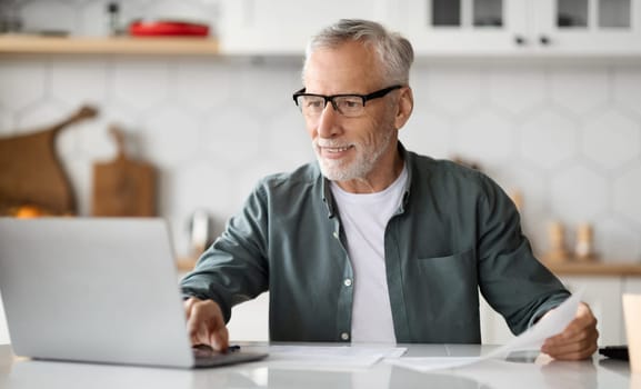 Business After Retirement. Smiling Senior Man Working With Laptop In Kitchen Interior