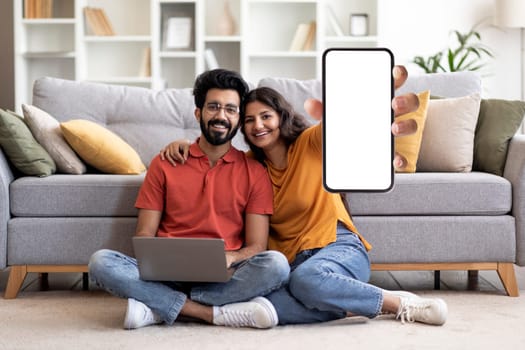 Happy Young Indian Couple Showing Blank Smartphone At Camera While Relaxing With Laptop At Home, Smiling Eastern Spouses Recommending New Mobile App Or Website, Creative Collage, Mockup