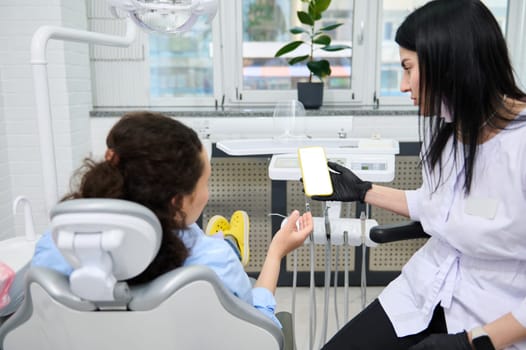 Rear view female dentist doctor holding smartphone with white blank digital mockup screen, copy ad space, and a patient at dental appointment in dentistry clinic. Dental practice. Visiting dentist