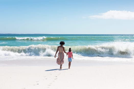 African american mother and daughter holding hands while walking towards waves at beach