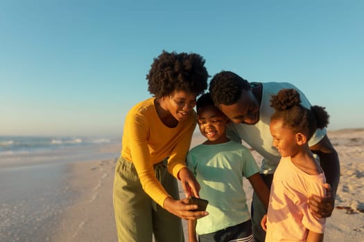 Happy african american mother sharing smartphone with family at beach on sunny day