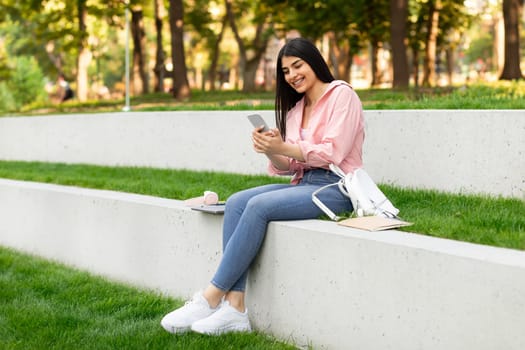 Smiling hispanic lady messaging on cellphone while relaxing in park or college campus outdoors, free space