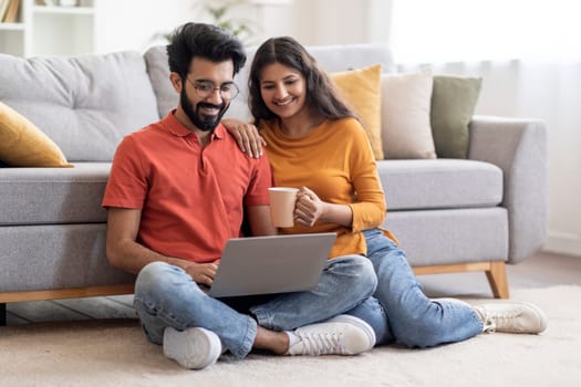 Smiling Indian Couple With Laptop And Coffee Relaxing In Living Room At Home, Happy Young Eastern Spouses Resting On Floor And Using Computer Together, Shopping Online Or Browsing Internet