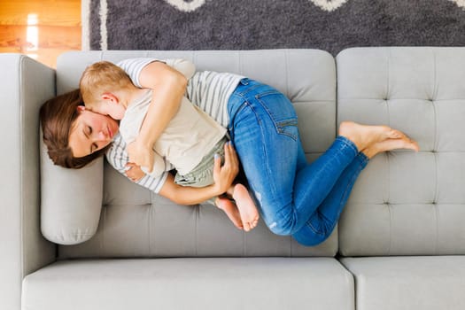Mother embraces her cute little son while laying together on the couch in the living room