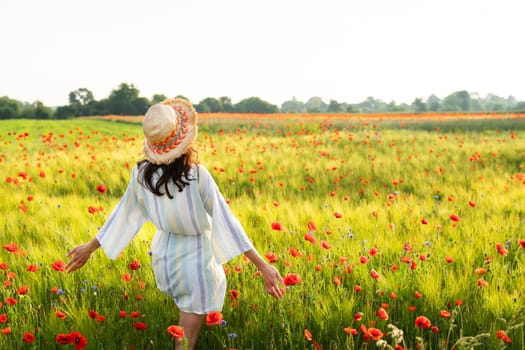 A girl in a straw hat, dress stands in a beautiful poppy field with red poppies. Sunrise, happiness, morning walk.