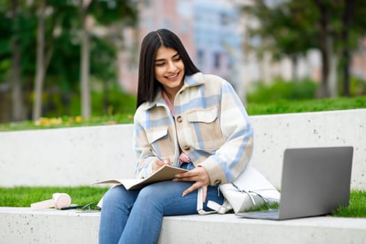 E-learning concept. Cheerful teen student girl learning outdoors, taking notes while sitting with laptop in park