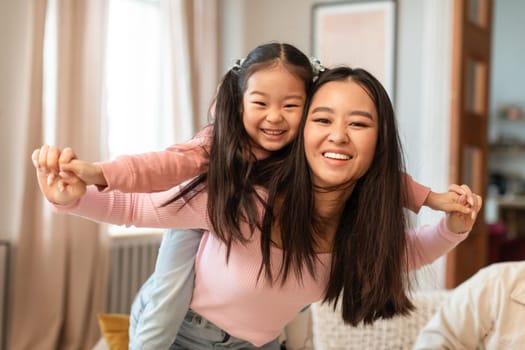 Family Playtime. Cheerful Asian Mom Carrying Little Daughter Piggyback Smiling At Camera, Posing Having Fun And Bonding At Home. Mommy And Child Playing Enjoying Weekend Leisure Indoor