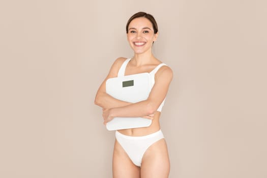 Thrilled young lady embracing electronic scales, beige background