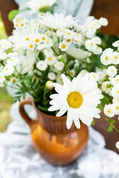 Beautiful chamomile flowers on a wooden old chair along with a lace doily on a green garden background. Summer atmosphere, simple home decor in the countryside. Vertical photo.
