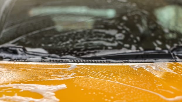 Closeup detail - front hood of yellow car covered with white shampoo foam when washed in carwash. Abstract automotive cleaning background with space for text.