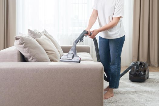 young woman in white shirt and jeans cleaning carpet under sofa with vacuum cleaner in living room, copy space. Housework, cleanig and chores concept