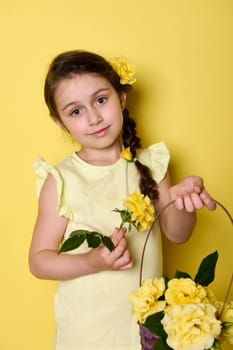 Beautiful little child girl holds wicker basket with yellow rose flowers, smiles looking at camera, isolated on yellow