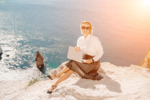 Business woman on nature in white shirt and black skirt. She works with an iPad in the open air with a beautiful view of the sea. The concept of remote work.