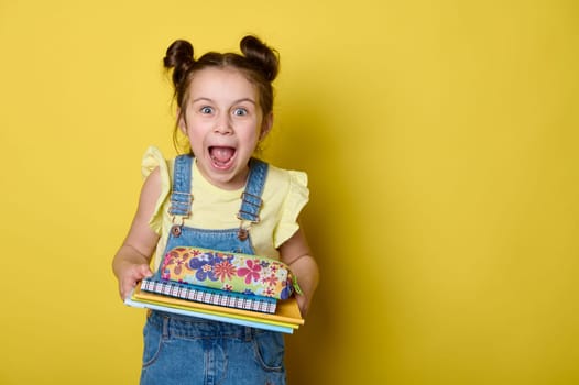 Cheerful kid girl with funny hairstyle, expressing amazement at camera, carrying books and pencil case, yellow backdrop