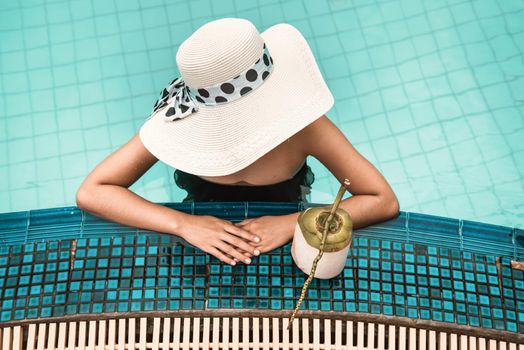Sexy Woman in Swimsuit is Relaxing in Swimming Pool, Beautiful Woman Wearing Straw Hat and Relax Sunbathe in Poolside on Summer Holiday at Resort Hotel. Summer Vacation and Relaxation Lifestyles