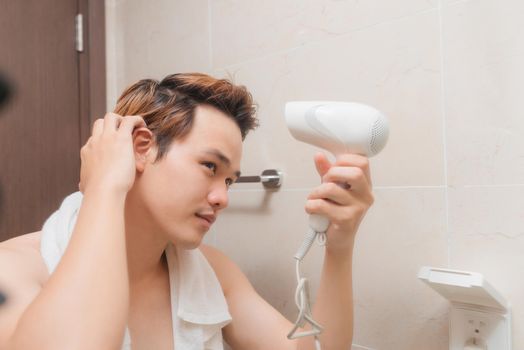 Trendy young man drying his hair with blow dryer in front of bathroom mirror