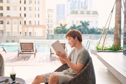  young man lying on the chaise-lounge and reading a book near the swimming pool