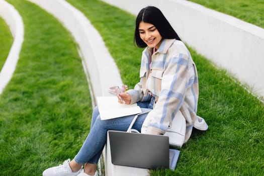 Happy lady student studying outdoors with laptop, having remote lesson and taking notes to notepad