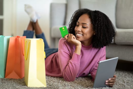 Happy millennial black woman lies on floor with packages with purchases, use tablet