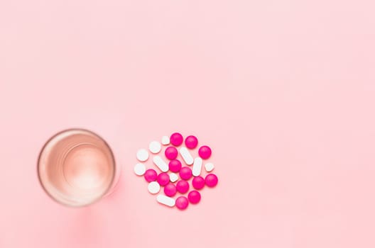 A glass of drinking water with piles of medicine pills on pink background for healthcare concept.