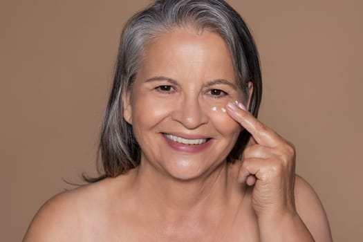 Positive european senior grey-haired woman enjoy daily procedure, applying cream on face