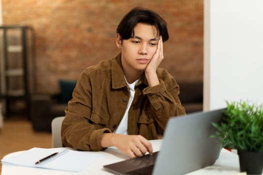 Bored Asian Student Guy Sitting Looking At Laptop At Home
