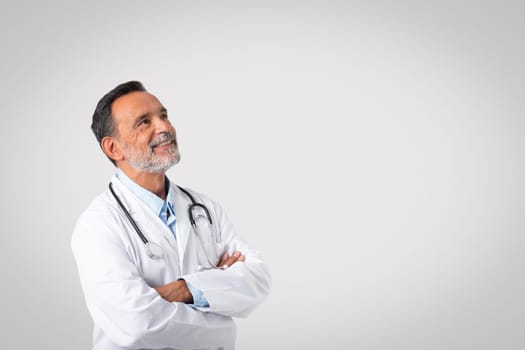 Glad confident caucasian mature doctor in white coat with crossed arms looks at copy space