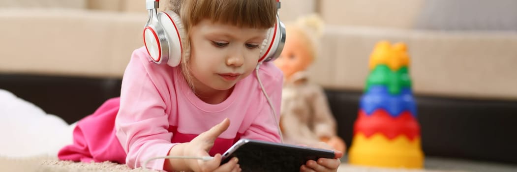 Little girl in headphones holds smartphone lying on floor