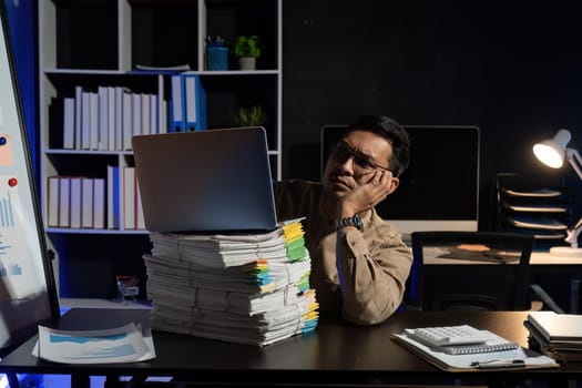 Overworked young Asian office employee working on laptop computer overtime in office at night