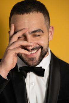 Shy looking down with eye covered with one hand handsome bearded man in tuxedo hide peeps out on closed eyes. Handsome young smiling caucasian man isolated on yellow background