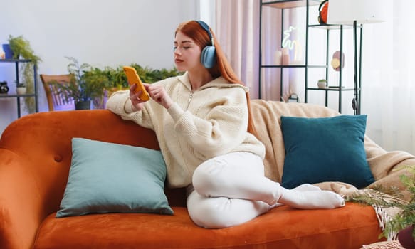 Happy overjoyed redhead woman in wireless headphones relaxing sits on couch at home apartment choosing listening favorite energetic disco dancing music. People weekends daytime leisure activities