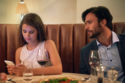 Phone, chatting and couple on a date at a restaurant for valentines day, romance or anniversary. Communication, upset and annoyed man watching his girlfriend sitting on her cellphone at dinner