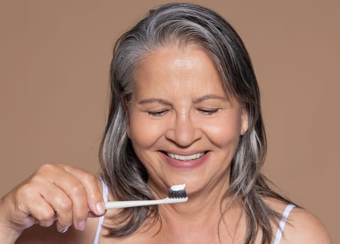Smiling european senior grey-haired woman brushing her teeth with toothbrush, enjoy daily routines