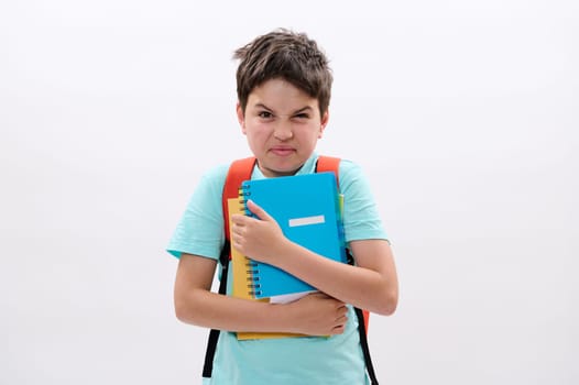 Irritated angry schoolboy holding workbooks, expresses negative emotions, feels unhappy to start new semester at school