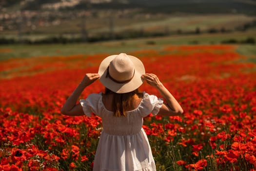 Field of poppies woman. Happy woman in a wight dress and hat stand through a blooming field of poppy. Standing with her back raised her hands up. Field of blooming poppies.