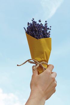 Close up woman hand holding bouquet of dried lavender flowers wrapped in yellow paper, raised up over blue sky, personal perspective, low angle view