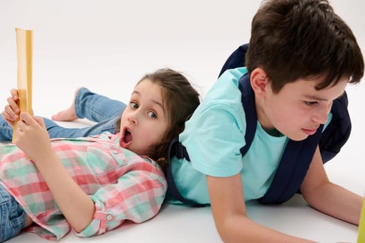 Preschooler girl or first grader with WOW emotion, looking at camera, lying next to her brother with a workbook in hands