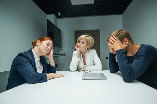 Blond, red-haired woman and bearded man in suits in the office.Colleagues sit at a table in a conference room and hold their heads in thought.