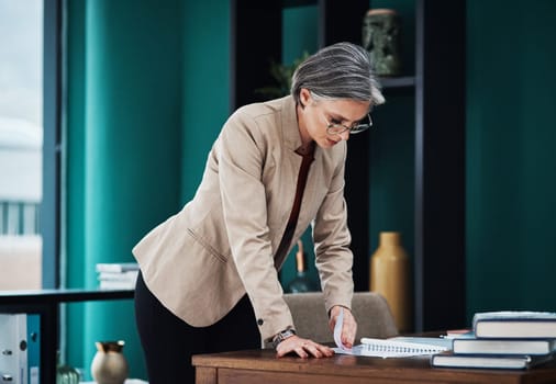The paperwork never ends. an attractive mature businesswoman standing and reading paperwork by her desk in her home office.