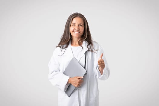 Professional approve service. Smiling senior european woman doctor in white coat showing thumb up, light background