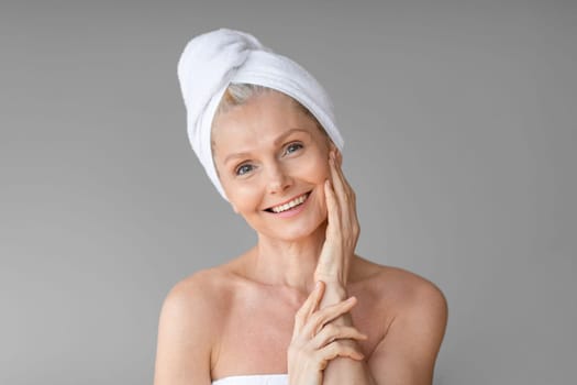 After spa. Portrait of happy aged woman with towel on head looking and smiling at camera, posing over grey background after beauty treatments, copy space