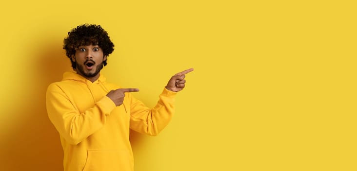 Amazed handsome eastern guy pointing at copy space, yellow background