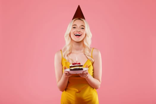 Portrait of excited lady wearing party hat, celebrating birthday, holding piece of cake with candle over pink background