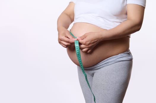 Gravid pregnant woman measuring her big tummy in the third trimester with a tape, isolated over white studio background