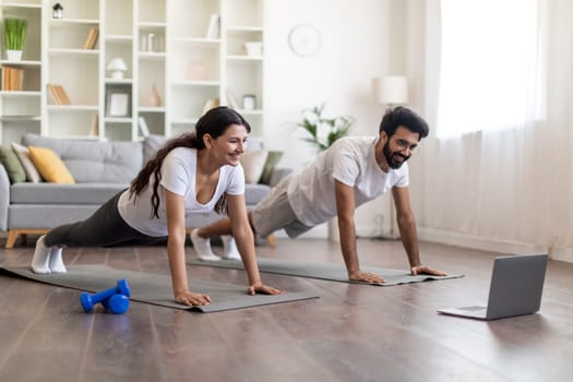 Sporty young indian couple in sportswear planking together in living room, happy eastern spouses having online fitness class or video call with trainer from home, using laptop computer, copy space