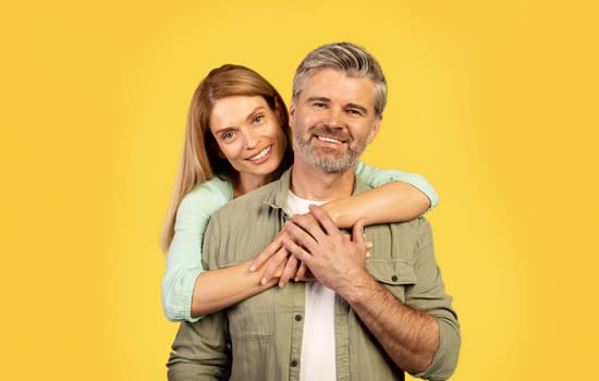 Portrait of happy middle aged woman hugging her husband from behind, posing on yellow background and smiling at camera